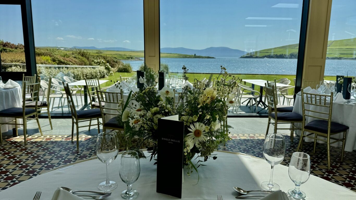 A set dining table with flower arrangements is positioned by large windows overlooking a scenic lake and hills under a clear blue sky.